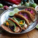 Three colorful tacos filled with ingredients like shrimp, avocado, and pickled onions, surrounded by fresh ingredients o