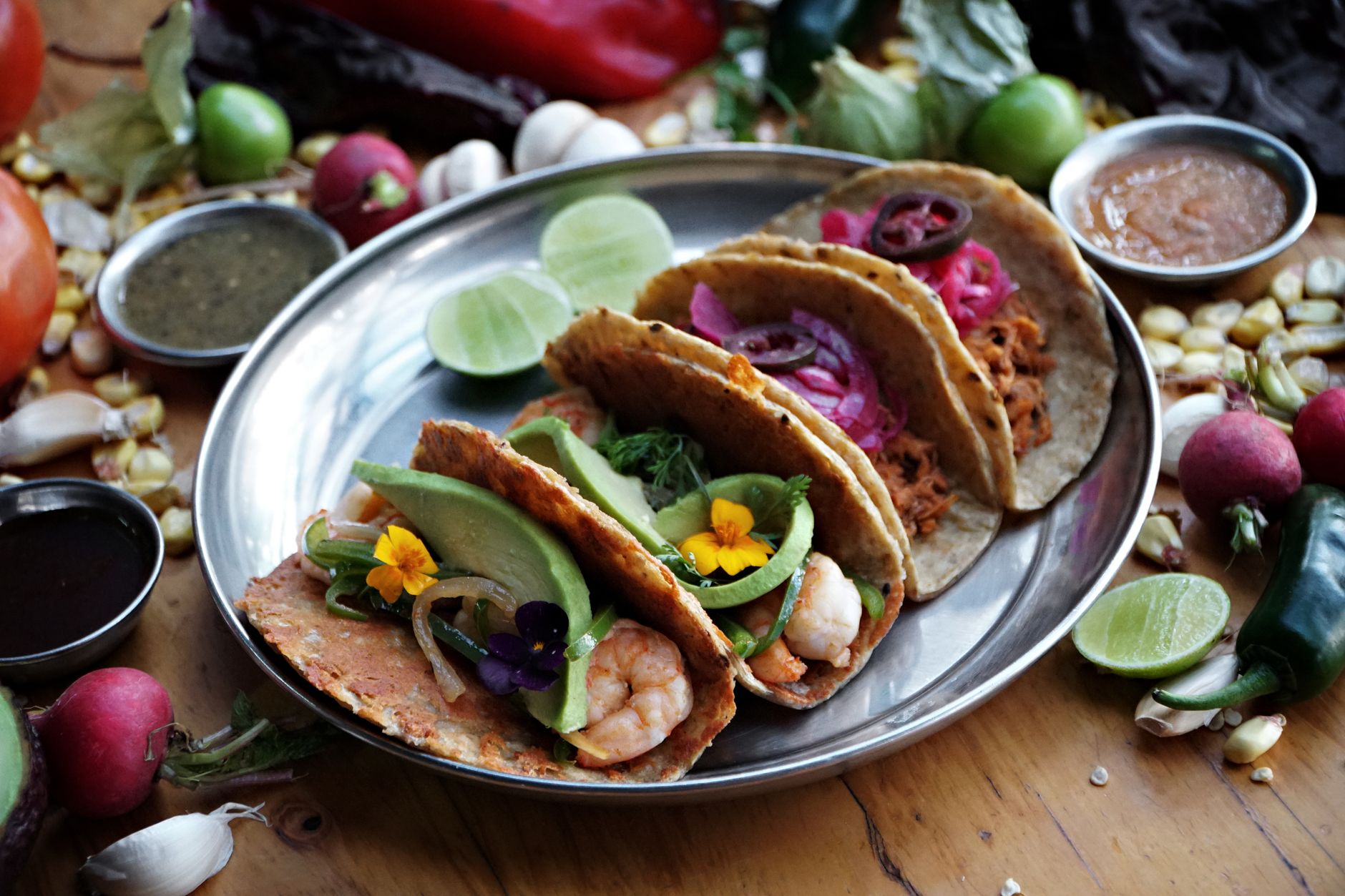 Three colorful tacos filled with ingredients like shrimp, avocado, and pickled onions, surrounded by fresh ingredients o